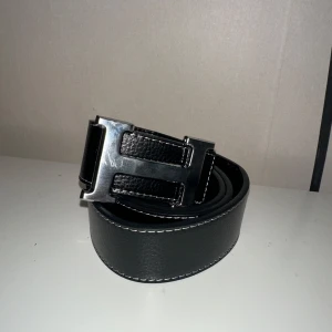 Svart bälte med metallspänne - Snyggt svart her-més bälte med ett stort, blankt metallspänne och vita kontrastsömmar längs kanterna. Bältet har en stilren design och är tillverkat i skinn. Perfekt accessoar för att lyfta din outfit. 100cm