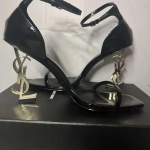 YSL-klackskor - Säljer ett par svarta sandaler från Saint Laurent med ikonisk YSL-klack i guld. Skorna har smal rem över tårna, ankelrem med spänne och fyrkantig tå. Tillverkade i glansigt skinn och levereras med originalpåse.