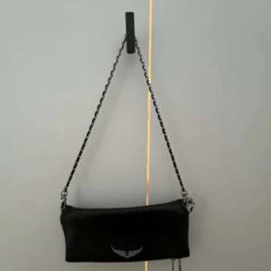 Svart axelväska med kedjerem - Säljer en svart axelväska med silverfärgad kedjerem och ett litet emblem framtill. Väskan har en avlång form och passar perfekt för att bära det viktigaste. Enkel och stilren design som funkar till många outfits.