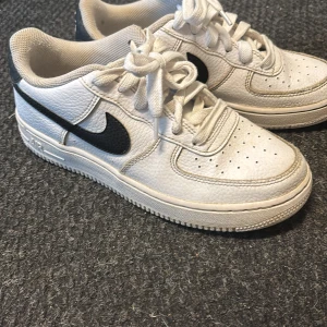 Nike Air Force 1 vita sneakers med svart swoosh - Klassiska Nike Air Force 1 sneakers i vitt skinn med svart swoosh på sidorna. Skorna har snörning, rund tå och platt sula. Perfekta för en stilren och sportig look.