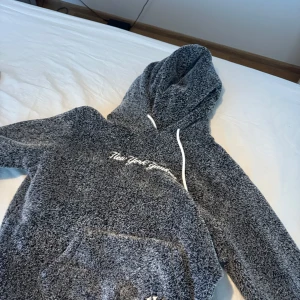 Grå fluffig hoodie med text - Mysig grå hoodie med fluffigt material och vit broderad text på bröstet. Tröjan har huva med vita snören och en stor magficka framtill. Perfekt för kalla dagar när du vill vara extra bekväm.