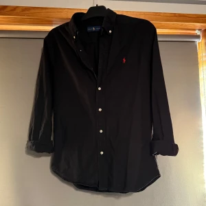 Svart skjorta från Ralph Lauren - Svart skjorta från Ralph Lauren med klassisk krage och vita knappar. Liten röd broderad logga på bröstet. Långärmad modell i bomull, perfekt för en stilren look.