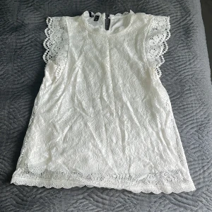 Vit spetsblus med korta ärmar - Säljer en söt vit blus i spets med fina virkade detaljer vid ärmslut och hals. Blusen har korta ärmar och knäppning bak i nacken. Perfekt att styla till jeans eller kjol!