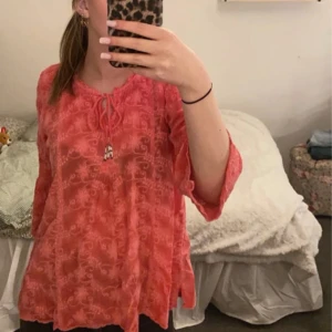 Korallröd blus med spetsdetaljer - Säljer en korallröd/rosa blus med fina spetsdetaljer och knytning vid halsen. Blusen har trekvartsärm och ett lätt, luftigt material som passar perfekt till våren och sommaren!