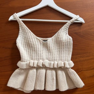 Beige stickad linnetopp med volang - Superfin beige stickad linnetopp med bred volangkant nedtill och ribbad struktur. Toppen har smala axelband och en söt, feminin look. Perfekt att styla till jeans eller kjol för en trendig outfit.