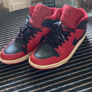 Nike Air Jordan 1 High röd och svart - Säljer ett par klassiska Nike Air Jordan 1 High i rött och svart med svarta skosnören och vit sula. Skorna har högt skaft, ikonisk vinge-logga på sidan och är perfekta för dig som gillar streetwear och sneakerskultur. Använts 1-2 gånger!