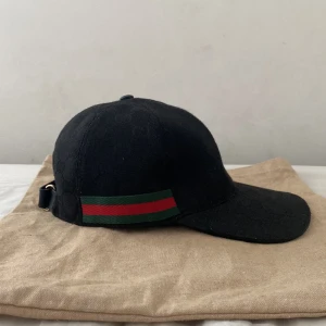 Svart keps från Gucci med gröna och röda detaljer - Säljer en svart keps från Gucci med klassiska gröna och röda ränder på sidorna. Kepsen har böjd skärm och justerbar rem baktill. Materialet är bomull med diskret GG-mönster i tyget. Perfekt för dig som gillar exklusiva accessoarer.
