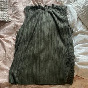 Olivgrön ärmlös blus med volangkant - Säljer en olivgrön ärmlös blus med smockad volangkant vid halsen. Blusen har ett subtilt randigt mönster och är tillverkad i ett lätt, mjukt tyg som faller snyggt. Perfekt för dig som gillar stilrena och bekväma plagg.