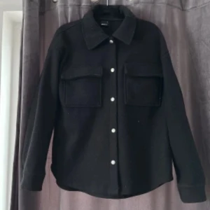 Svart overshirt med fickor - Snygg svart overshirt med klassisk krage och två stora bröstfickor. Skjortan har långa ärmar och knäpps med silverfärgade knappar framtill. Jätte skön att ha på sig och fin. Mer bilder skiv !