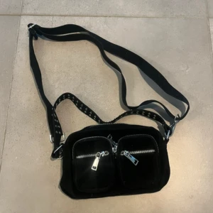 Svart axelväska  - Säljer en svart axelväska med silvriga detaljer. Väskan har tillhörande långt och kort axelband. Den är i nyskick och oanvänd!!