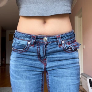 Blå bootcut jeans med röda sömmar från True Religion - Säljer ett par blå bootcut jeans från True Religion, modell BECCA Mid Rise Bootcut. Jeansen har röda kontrastsömmar, klassiska fickor bak med lock och knapp samt normal passform. Perfekta för dig som gillar detaljer och unik stil. Size: 26. Waist:35-36