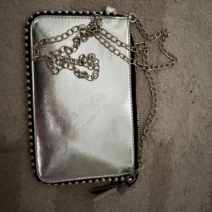 Silver axelväska med kedja - Säljer en snygg axelväska i metallic silver med kedjerem och dekorativa nitar längs kanterna. Väskan har dragkedja upptill och är perfekt för att lyfta din outfit med en cool detalj.