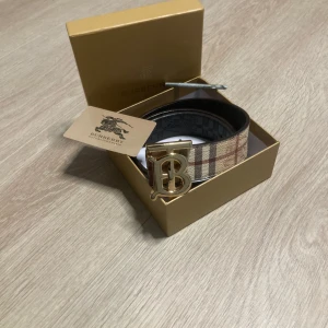 Burberry bälte med guldspänne - Säljer ett Burberry bälte med klassiskt rutigt mönster i beige, rött och svart. Bältet har ett stort guldigt spänne med Burberrys logotyp. Box kommer ej med
