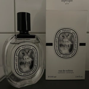 L'Eau Papier Eau de Toilette från Diptyque - Ca 95 ml kvar. En elegant parfym i en genomskinlig oval glasflaska med svart lock. Märket Diptyque och doftnamnet L'Eau Papier syns tydligt på etiketten. Flaskan rymmer 100 ml och har en stilren, konstnärlig design med svartvit etikett. 