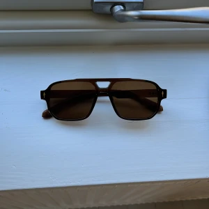 Bruna solglasögon med fyrkantig form - Snygga solglasögon med bruna glas och markerad fyrkantig båge i mörkbrunt. Bågarna har en klassisk design med raka linjer och detaljer i guld vid gångjärnen. Perfekta för en stilren look.