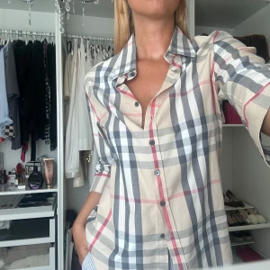 Beige rutig skjorta från Burberry - Säljer en klassisk beige skjorta från Burberry med ikoniskt rutigt mönster i svart, vitt och rött. Skjortan har lång ärm, knappar framtill och en avslappnad passform. Är i storlek L men passa alla från XS till M beroende på hur oversized man vill ha, endast använt ett fåtal gånger och är i mycket bra skick.