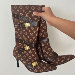 Bruna stövletter med monogram och gulddetaljer - Säljer ett par bruna stövletter med klassiskt monogrammönster och svarta läderremmar med guldfärgade spännen. Skorna har spetsig tå, smal klack och dragkedja på insidan. Tidigare köpta på en second hand butik.