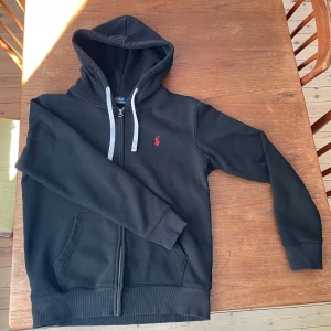 Svart hoodie från Polo Ralph Lauren - Säljer en svart hoodie från Polo Ralph Lauren med dragkedja och vit snörning i huvan. Klassisk röd broderad logga på bröstet och två fickor framtill. Perfekt för en avslappnad stil. Den har dock en lagning i en av framfickorna, se sista bilden!