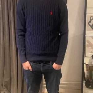 Mörkblå kabelstickad tröja från Ralph Lauren L - Säljer en mörkblå kabelstickad tröja från Ralph Lauren med röd broderad logga på bröstet. Tröjan har rund halsringning och långa ärmar. Klassisk och stilren modell som passar till mycket.