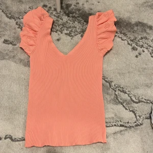 Rosa ribbad topp med volangärmar - Superfin rosa topp med ribbad struktur och v-ringning. Ärmarna har volangdetaljer som ger en söt och trendig look. Perfekt för vår och sommar!