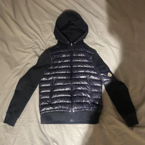 🧥 Moncler Maglia Tricot Cardigan – Marinblå / Storlek S - Säljer en äkta Moncler cardigan i stilren marinblå färg. Jackan kombinerar en lätt vadderad, glansig front med mjuka ärmar och rygg i bomull – perfekt för både vardag och lager-på-lager stil.