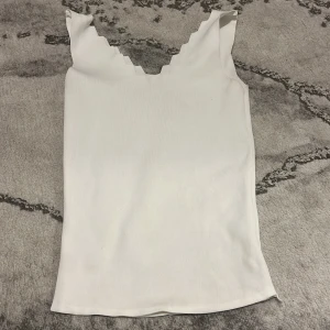 Vit ribbad linnetopp med vågig kant - Säljer en vit ribbad linnetopp med snygg vågig kant vid axlar och halsringning. Toppen är ärmlös och har en figurnära passform, perfekt att styla till olika outfits.