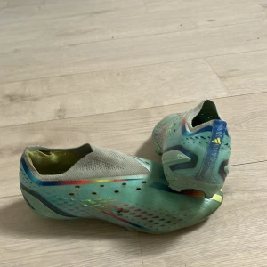 Adidas fotbollsskor i turkos och silver - Säljer ett par Adidas fotbollsskor i turkos med inslag av silver, blått och regnbågsfärgade detaljer. Skorna har låg profil, platt sula och är tillverkade i syntetmaterial. Perfekta för dig som vill sticka ut på planen!