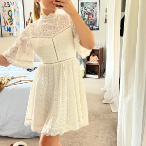 By Malina Klänning - Superfin vit klänning i spets med halvlånga, vida ärmar och hög hals. Klänningen har vackra broderade detaljer, knappar framtill och en lätt utställd kjol. Perfekt för sommarens alla tillfällen!