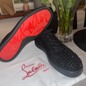 Svarta Louboutins - Säljer ett par svarta sneakers från Christian Louboutin med ikoniska röda sulor och svarta nitar på tån. Skorna har snörning och är tillverkade i skinn. Perfekta för dig som vill sticka ut med en lyxig och edgy stil. Är använda några gånger men annars inte så mycket. Mvh