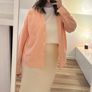 Randig skjorta från H&M - Säljer en skjorta från H&M med orange/korall och vita ränder. Skjortan har lång ärm, klassisk krage och bröstficka. Perfekt att styla såväl öppen som knäppt. Aldrig använd.