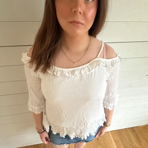 Vit offshoulder blus med volang - Säljer en söt vit offshoulder blus med volangdetaljer längs halsringningen och nederkanten. Blusen har trekvartsärm och är tillverkad i ett lätt, krinklat material som ger en avslappnad känsla.