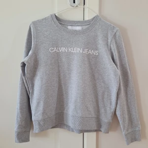 Grå sweatshirt från Calvin Klein Jeans - Säljer en klassisk grå sweatshirt från Calvin Klein Jeans med vit logotyp framtill. Tröjan har rund halsringning, långa ärmar och ribbade muddar vid ärmslut och nederkant. Passar mer strl S