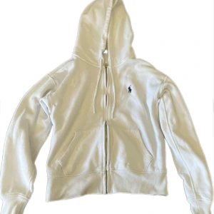 Vit hoodie från Polo Ralph Lauren - Säljer en klassisk vit hoodie från Polo Ralph Lauren med dragkedja och huva. Tröjan har två fickor framtill och en broderad logga på bröstet. Perfekt för en avslappnad och stilren look. Nypris 1600 kr mitt pris 699. Skicket är 10/10 inget fel på hoodien, är även öppen för frågor.