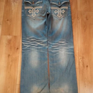 baggy olimp jeans - Säljer ett par blå bootcut jeans med feta broderade detaljer på bakfickorna och slitningar framtill. Liknar rock revival