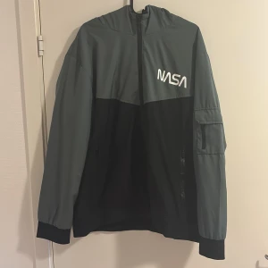 Svart och grå NASA vindjacka - Snygg vindjacka i svart och grått med NASA-tryck på bröstet. Jackan har huva, dragkedja framtill och en ficka på ärmen. Perfekt för dig som gillar rymdtema och streetwear.