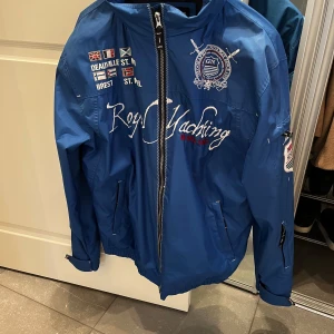 Blå vindjacka från Geographical Norway - Säljer en blå vindjacka från Geographical Norway med broderade detaljer och tryck både fram och bak. Jackan har dragkedja, ståkrage och flera fickor. Perfekt för dig som gillar en sportig och stilren look. Storlek L men sitter som M