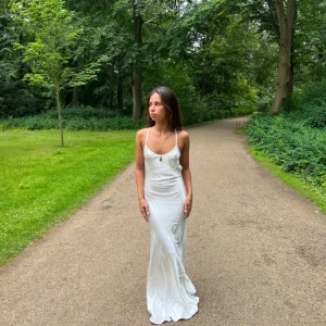 Vit långklänning med smala axelband - Säljer en elegant vit långklänning med smala axelband och öppen rygg. Klänningen har en mjuk, glansig yta och knytning i ryggen för en snygg passform. Perfekt för sommarens alla tillfället eller till balen 💕💕