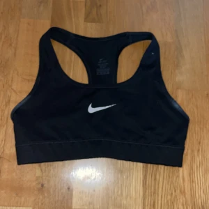 Svart sport-bh från Nike - Svart sport-bh från Nike med klassisk vit logga framtill. Modellen har brottarrygg och bred resår nedtill för extra stöd. Perfekt för träning och sportiga aktiviteter.