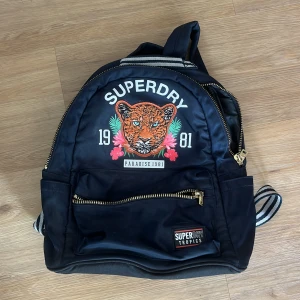 Superdry ryggsäck med leopardtryck - Cool svart ryggsäck från Superdry med stort leopardmotiv, tropiska blommor och texten 'Paradise 1981' framtill. Väskan har guldfärgade dragkedjor, randiga axelband och ett extra fack framtill. Perfekt för skolan eller stan.