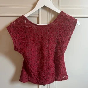 Vinröd spetsblus från Stradivarius - En sparsamt använd supersnygg vinröd blus från Stradivarius i spets med korta ärmar och öppen rygg. Blusen har ett vackert spetsmönster och en lätt croppad passform. Passar storlek XS-M, men står storlek L