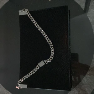 Svart kuvertväska med kedja - Snygg svart kuvertväska i skinnimitation med reptilmönstrad yta och en silverfärgad kedja som detalj. Väskan har ett rymligt fack och innerficka med dragkedja. Den är oanvänd .