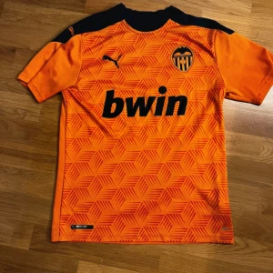 Valencia CF orange matchtröja Puma S - Säljer en Valencia CF matchtröja från Puma i orange med svarta detaljer. Tröjan har korta ärmar, geometriskt mönster framtill och klubbmärke på bröstet. Materialet är lätt och andas, perfekt för fotboll eller träning. Sponsorlogga 'bwin' tryckt på framsidan.