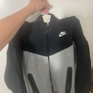 Nike tech svart/grå XS - Nike tech med dragkedja i svart och grått. Snygg tvåfärgad design med vit Nike-logga på bröstet och praktiska fickor framtill. Mjuk och skön, perfekt för chill eller träning. Storlek XS.