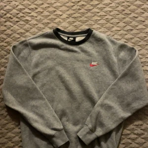 Grå Nike sweatshirt med röd vit logo - Grå sweatshirt från Nike med klassisk broderad logga i vitt och rött på bröstet. Tröjan har rund halsringning med svart kant, långa ärmar och ribbade muddar. Perfekt för en chill och sportig stil.