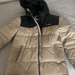 Beige och svart pufferjacka Everest - Snygg pufferjacka från Everest i beige med svarta axelpartier och huva. Jackan har dragkedja framtill, två fickor med dragkedja och quiltad design. Perfekt för kalla dagar när du vill hålla stilen och värmen.