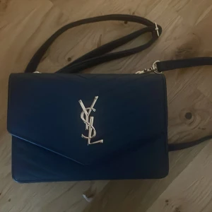 Mörkblå YSL axelväska i skinn - Säljer en mörkblå axelväska från YSL med klassisk silverfärgad logga framtill. Väskan är tillverkad i skinn och har quiltad design, justerbar axelrem och dragkedja upptill. Perfekt storlek för det viktigaste och ger en lyxig vibe till din outfit. Jag har aldrig använt den.
