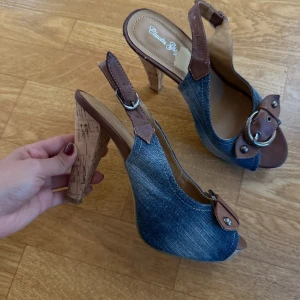 Jeansklackar med bruna detaljer - Unika klackar i jeansmaterial med bruna skinnremmar och stora silverspännen framtill. Snygg korkklack och öppen tå ger en cool vibe. Perfekta för dig som vill sticka ut med en mix av denim och läder.
