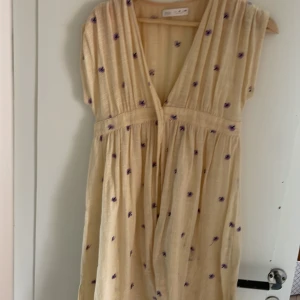 Ljusgul blommig klänning från Zara - Supersöt gul klänning från Zara med lila och blå broderade blommor. Djup v-ringning, korta ärmar och knappar framtill. Tillverkad i tunnt luftigt tyg perfekt för varma dagar. Snyggt rynkad midja ger en avslappnad och flowy känsla.