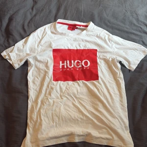 Vit t-shirt från Hugo Boss med röd logga - Vit t-shirt från Hugo Boss med stor röd logga och texten HUGO på bröstet. Klassisk passform med rund hals och korta ärmar. Tillverkad i mjuk bomull, perfekt för en clean och stilren look. Röd detalj på insidan av kragen för extra edge. Storlek M