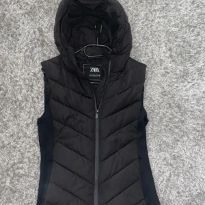 Svart dunväst med huva från Zara - Snygg svart dunväst från Zara med huva och dragkedja framtill. Västen har quiltad design med sneda sömmar och en figurnära passform. Perfekt att slänga över en hoodie eller tröja när det är kyligt ute.